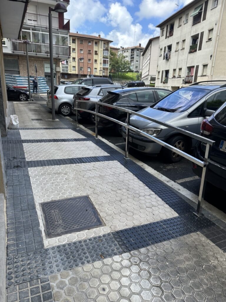 Acceso peatonal con barandilla en una calle con vehículos estacionados