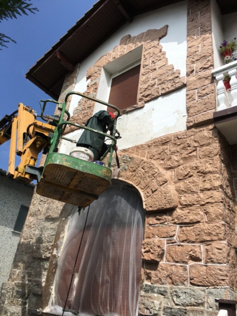 Trabajador en canasta elevadora realizando reformas en una fachada de piedra color beige