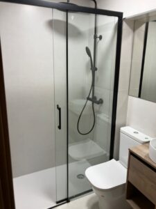 Ducha con mampara de vidrio en un baño nuevo, revestido en tonalidades claras.