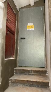 Puerta metálica gris con manija y cartel de aviso, escalones de acceso en la escena