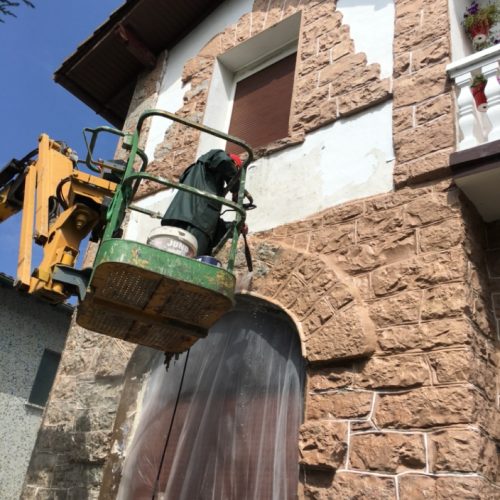 Trabajador en canasta elevadora realizando reformas en una fachada de piedra color beige